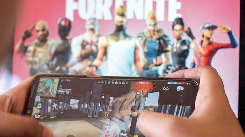 Ein Kind spielt ein Spiel auf einem Mobiltelefon mit einem Bild des Fortnite-Spiels auf dem Computerbildschirm im Hintergrund. (Archivbild). Foto: Herwin Bahar/ZUMA Wire/dpa