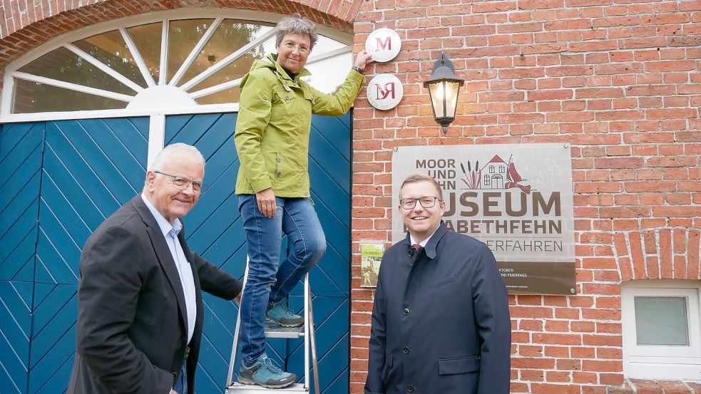 Museumsleiterin Antje Hoffmann ließ es sich nicht nehmen, die Metallplakette selber an der Außenwand des Moor- und Fehnmuseums anzubringen. Hans Eveslage (links) und Nils Anhuth assisierten. Foto: Kruse