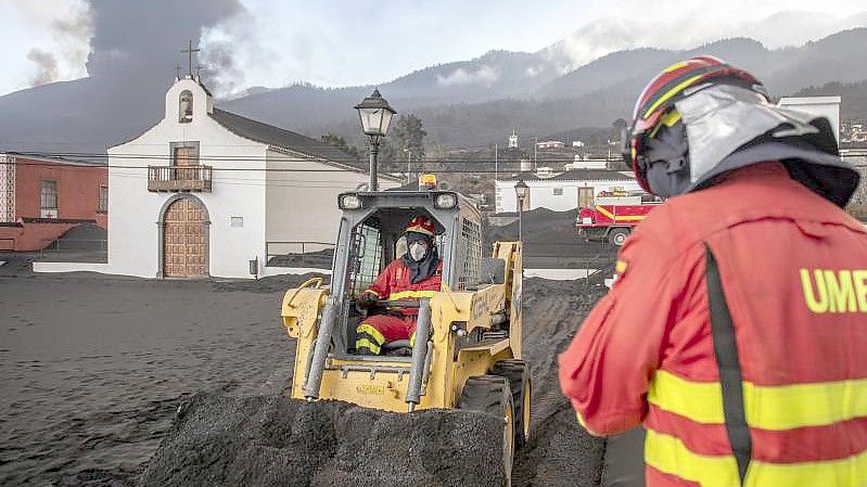 Einsatzkräfte des Militärs entfernen schwarze Asche vom Vulkan, der hinter der kleinen Kirche auf der Kanareninsel La Palma weiter Lava ausstößt. Foto: Saul Santos/AP/dpa