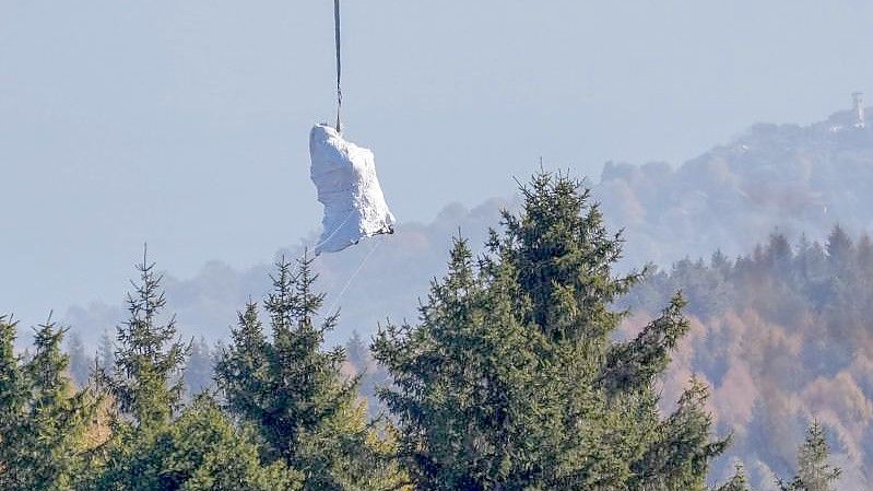 Ein Hubschrauber der italienischen Feuerwehr fliegt die in weiße Planen gehüllte Kabine der abgestürzten Mottarone-Seilbahngondel vom Lago Maggiore ins Tal. Foto: Antonio Calanni/AP/dpa