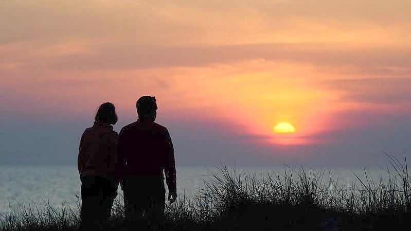 Ein Paar steht während des rötlich-orangenen Sonnenunterganges über der Ostsee an einer Düne am Strand. Foto: Gregor Fischer/dpa