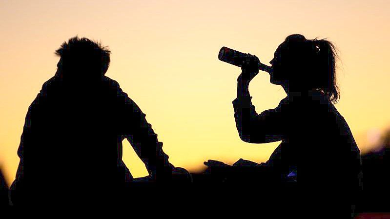 Ein Mann und eine Frau sitzen in der Abenddämmerung am Rheinufer und trinken aus Flaschen. (Archivbild). Foto: Marius Becker/dpa