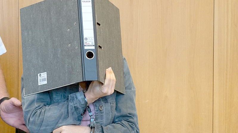 Die Angeklagte verdeckt ihr Gesicht mit einem Aktenordner, während sie in Handschellen in den Schwurgerichtssaal des Landgerichts in Ulm geführt wird. Foto: Stefan Puchner/dpa