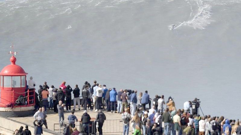 Menschen am Leuchtturm bei Nazaré beobachten einen Surfer am Praia do Norte, der eine Riesenwelle surft. Foto: Armando Franca/AP/dpa