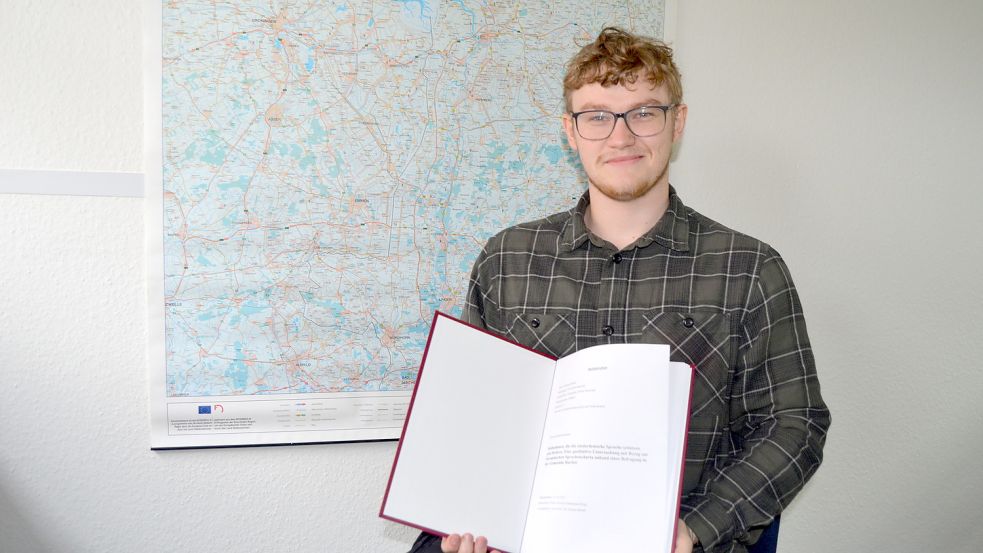 Der 23-jährige Barßeler Henning Schoone mit seiner Bachelorarbeit über das Plattdeutsche vor einer Landkarte Ostfrieslands, wo Platt immer noch sehr verbreitet ist. Foto: Fertig