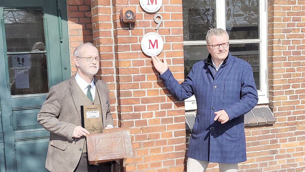 Gemeinsam mit Bürgermeister Theo Douwes hat Udo Tinnemeyer die neue Plakette zur Auszeichnung inzwischen an den Eingang des Schulmuseums angeschraubt. Tinnemeyer hält eine Entdeckertasche hoch. Foto: Gemeinde Westoverledingen