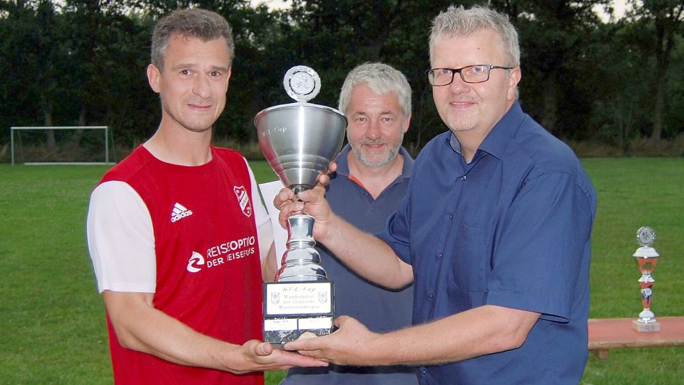 Ihrhoves Kapitän Paul Tomaszewski erhält den Pokal von Gemeindebürgermeister Theo Douwes (rechts) und dem Fresena-Vorsitzenden Ludwig Meinders. Foto: Reemts
