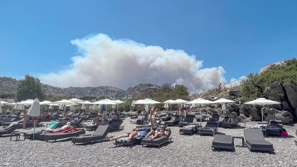 Am Viglika Beach auf der griechischen Insel Lindos aus, wo sich Menschen auf Sonnenliegen am Strand aufhalten, kann man aufsteigenden Rauch von Waldbränden auf der Ferieninsel Rhodos sehen. Foto: dpa/News Images via ZUMA Press Wire/Mark Cosgrove