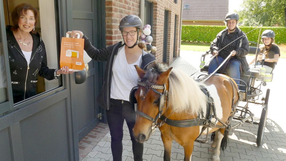 Käthe Gröne von der Apotheke am Denkmal in Flachsmeer (links) reicht Rebecka Müller aus dem Apothekerfenster eine Tüte mit Medikamenten. Müller führt Pony Joke. Auf dem Kutschbock sitzt Janina Robben und hinter ihr sitzt die zehnjährige Charlotte Müller. Foto: Wieking