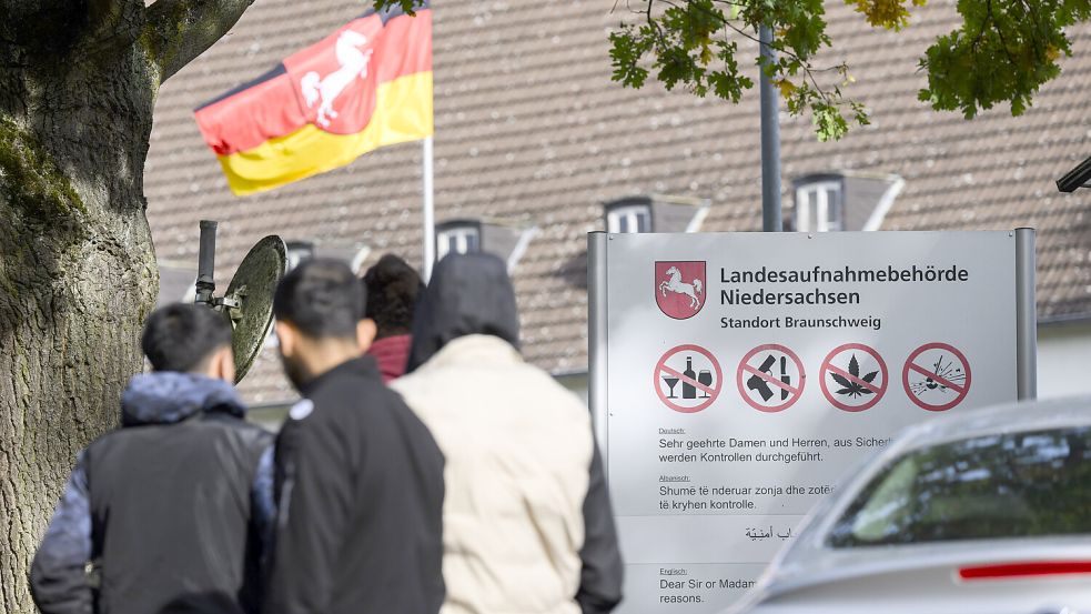 Männer stehen vor einem Schild der Landesaufnahmebehörde Niedersachsen am Standort Braunschweig. Die Zahl geflüchteter Menschen, die nach Niedersachsen kommen, wird auf absehbare Zeit nicht sinken. Bei der Unterbringung sind Land und Kommunen an ihre Grenzen gekommen. Foto: Julian Stratenschulte/dpa