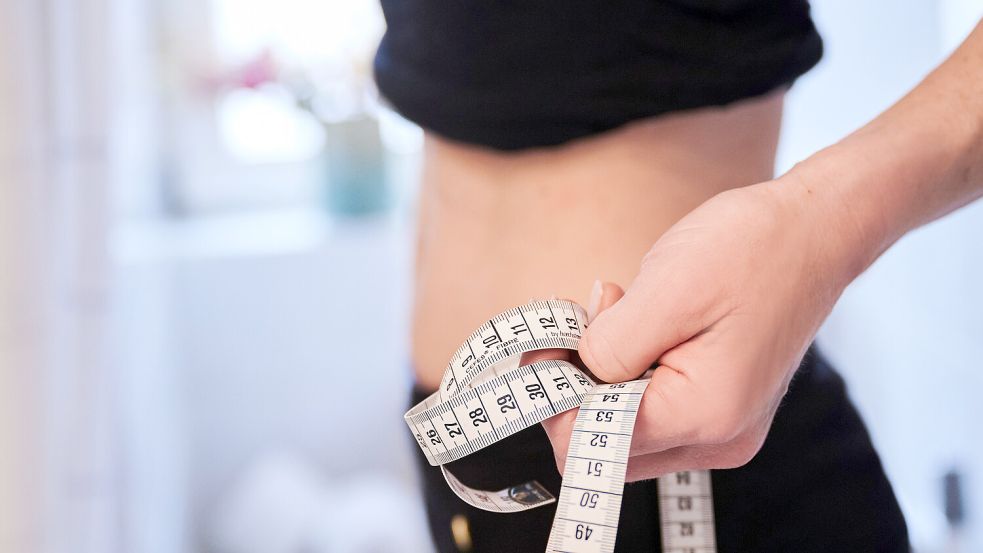 Eine junge Frau hält ein Zentimetermaßband an ihren Bauch. Ein übermäßiges Kontrollieren von Gewicht und Körpermaßen kann ein Anzeichen für eine Essstörung sein. Foto: Annette Riedl