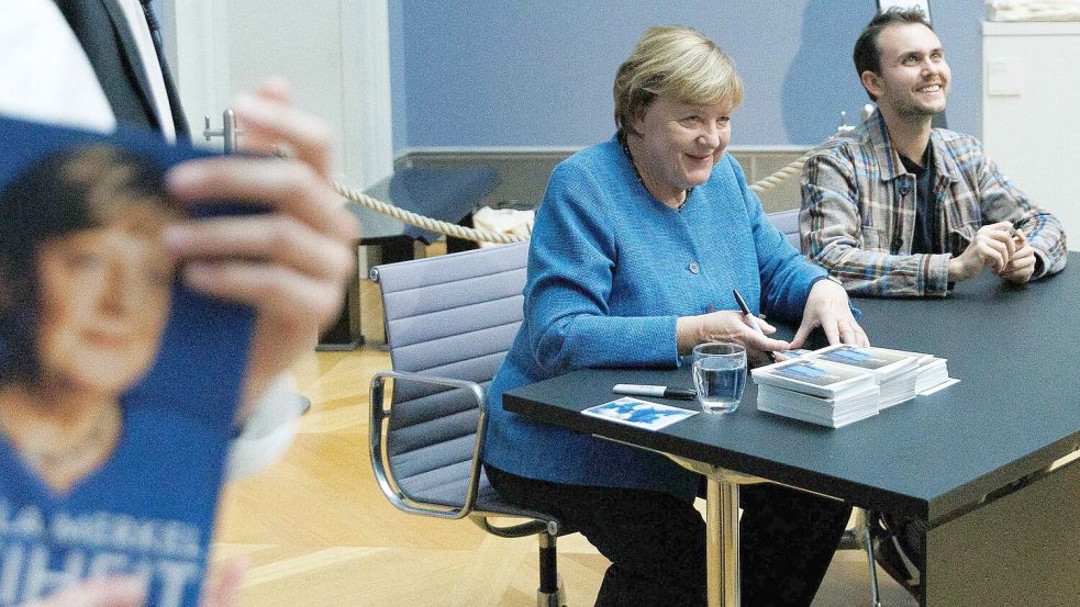 Merkel signiert Kunstpostkarten im Bode-Museum neben dem Maler ihres dort ausgestellten Porträts, Jérémie Queyras.  Foto: Carsten Koall/dpa